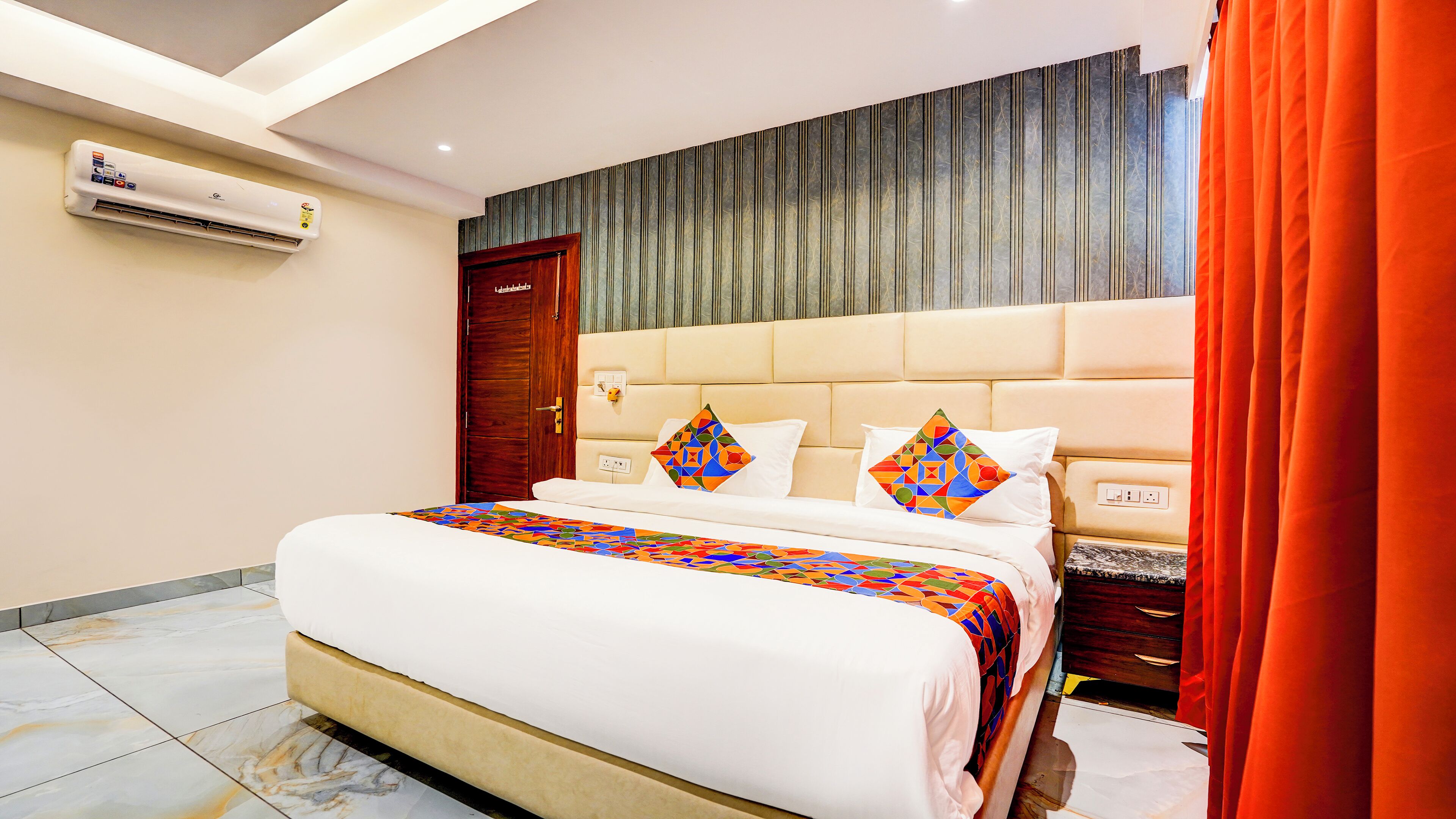 A hotel room with a large bed, a wooden door, an air conditioning unit, and a patterned curtain.