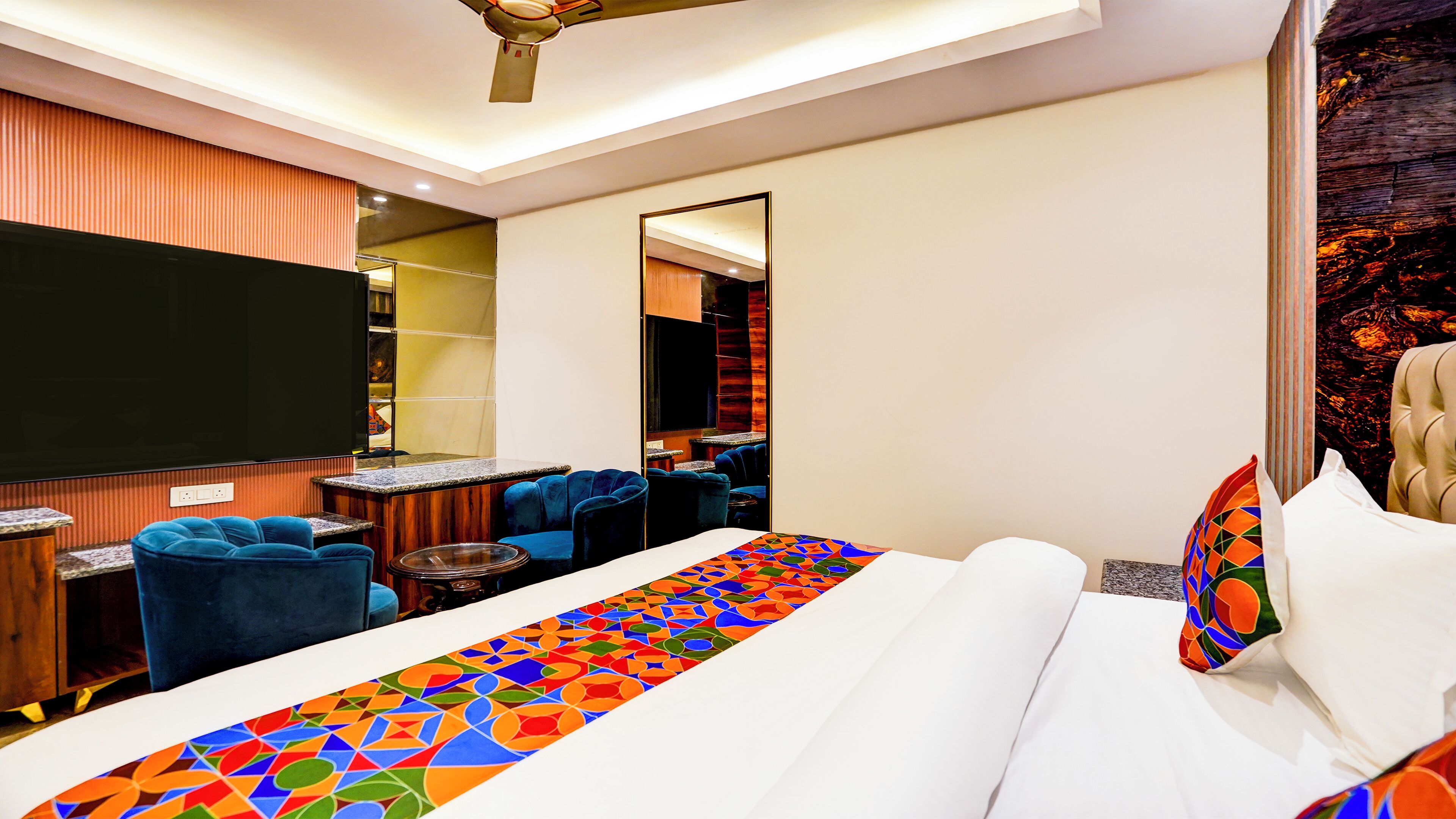 A hotel room with a large bed, a TV, a seating area with blue chairs, and a mirror.
