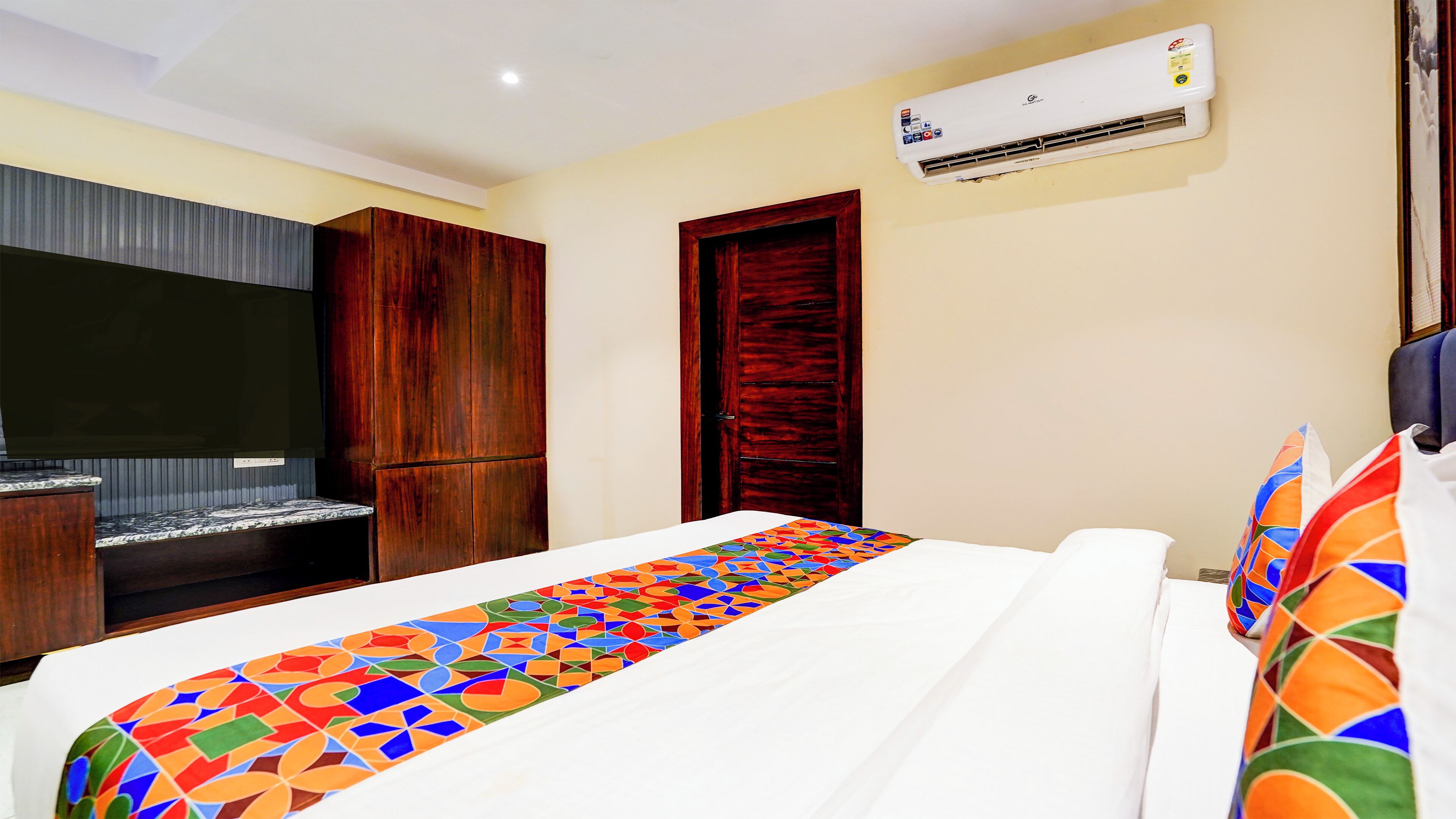 A hotel room with a bed, a TV, wooden wardrobe, a door, and an air conditioning unit.