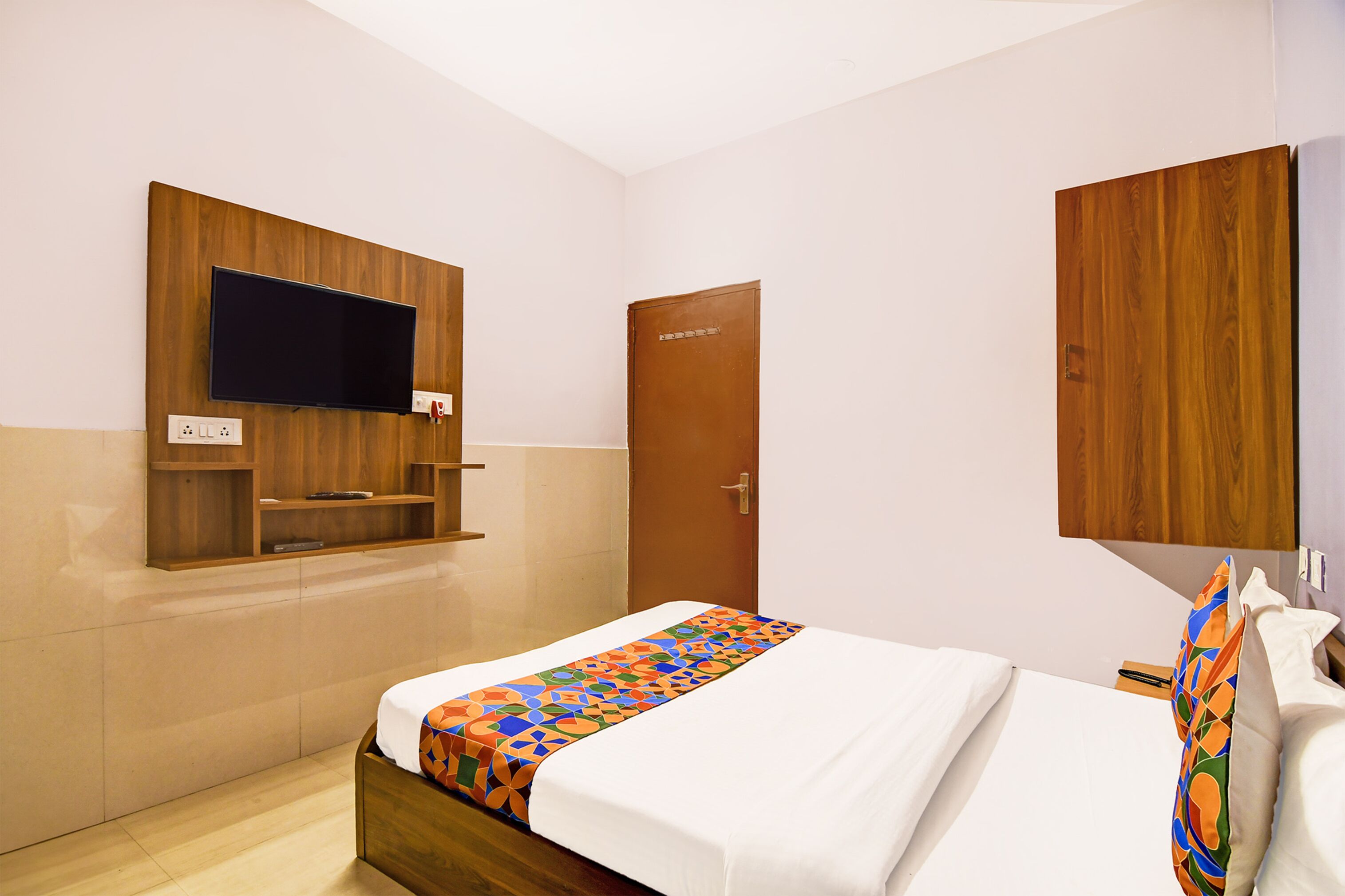 A hotel room with a bed, a TV mounted on the wall, a wooden door, and a wooden cabinet.