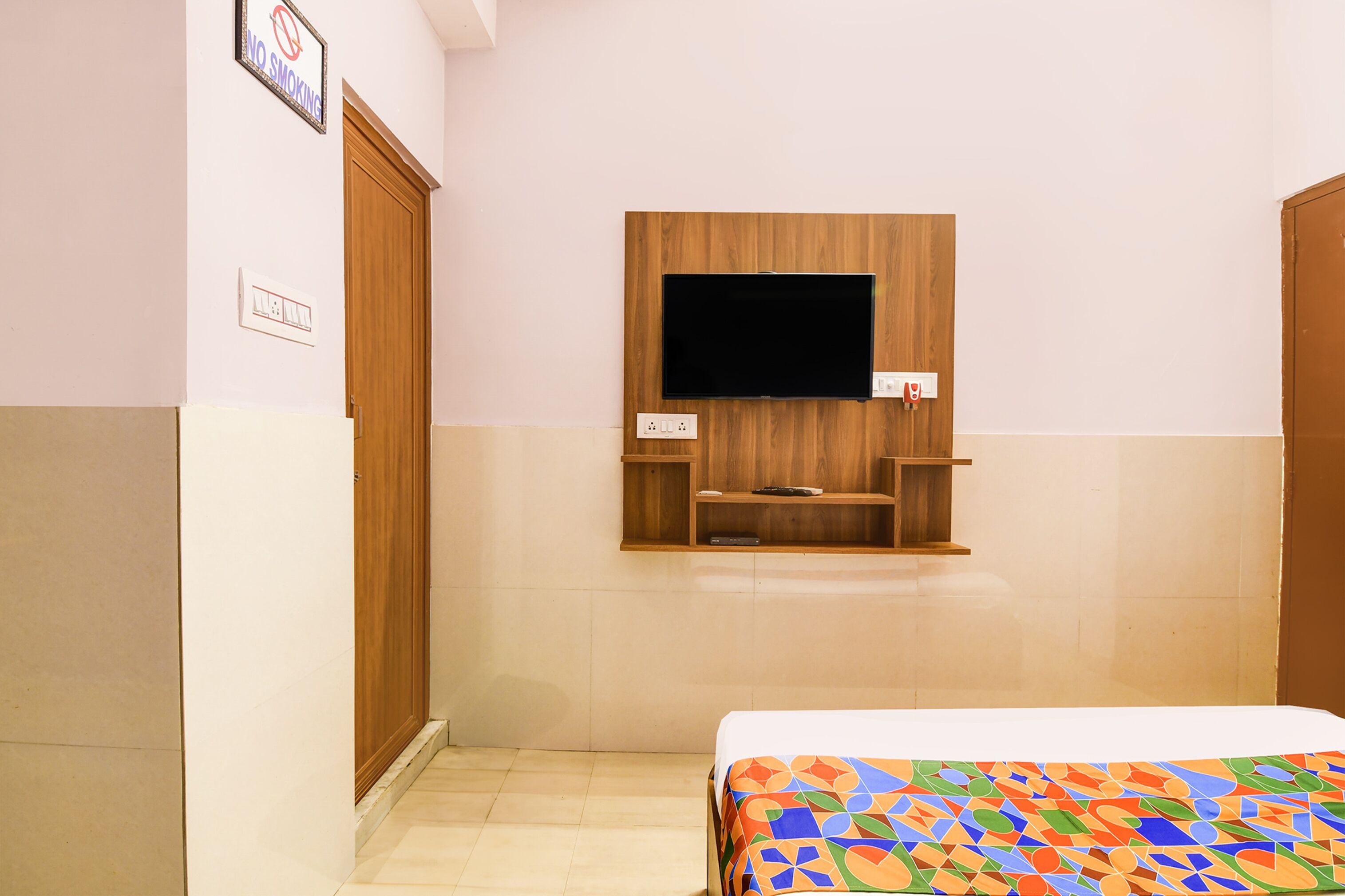 A hotel room with a bed, a TV mounted on a wooden panel, a small shelf, and a door.