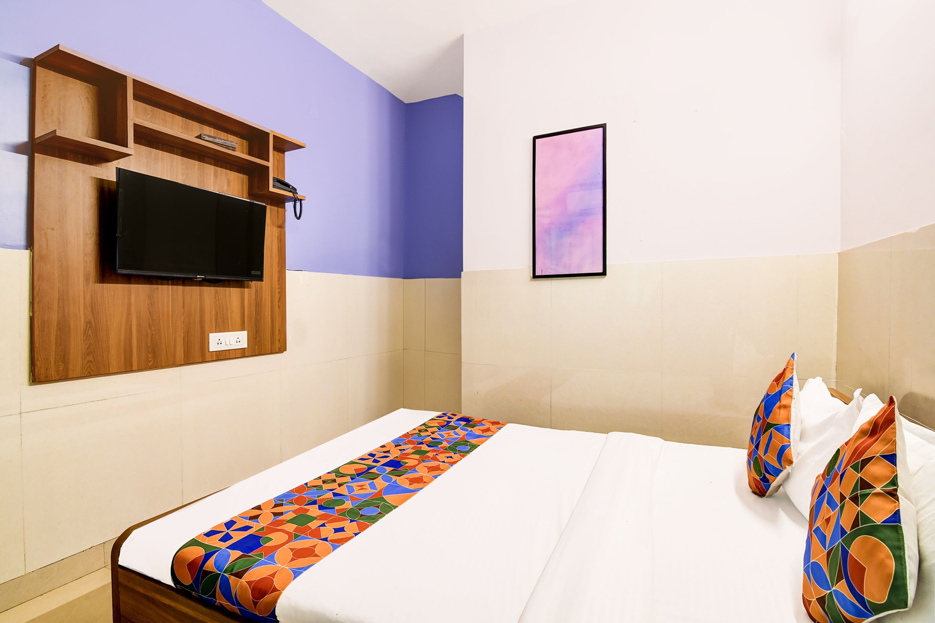 Una habitación de hotel con una cama, un televisor montado en la pared, un cuadro enmarco y un corredor de cama colorido.