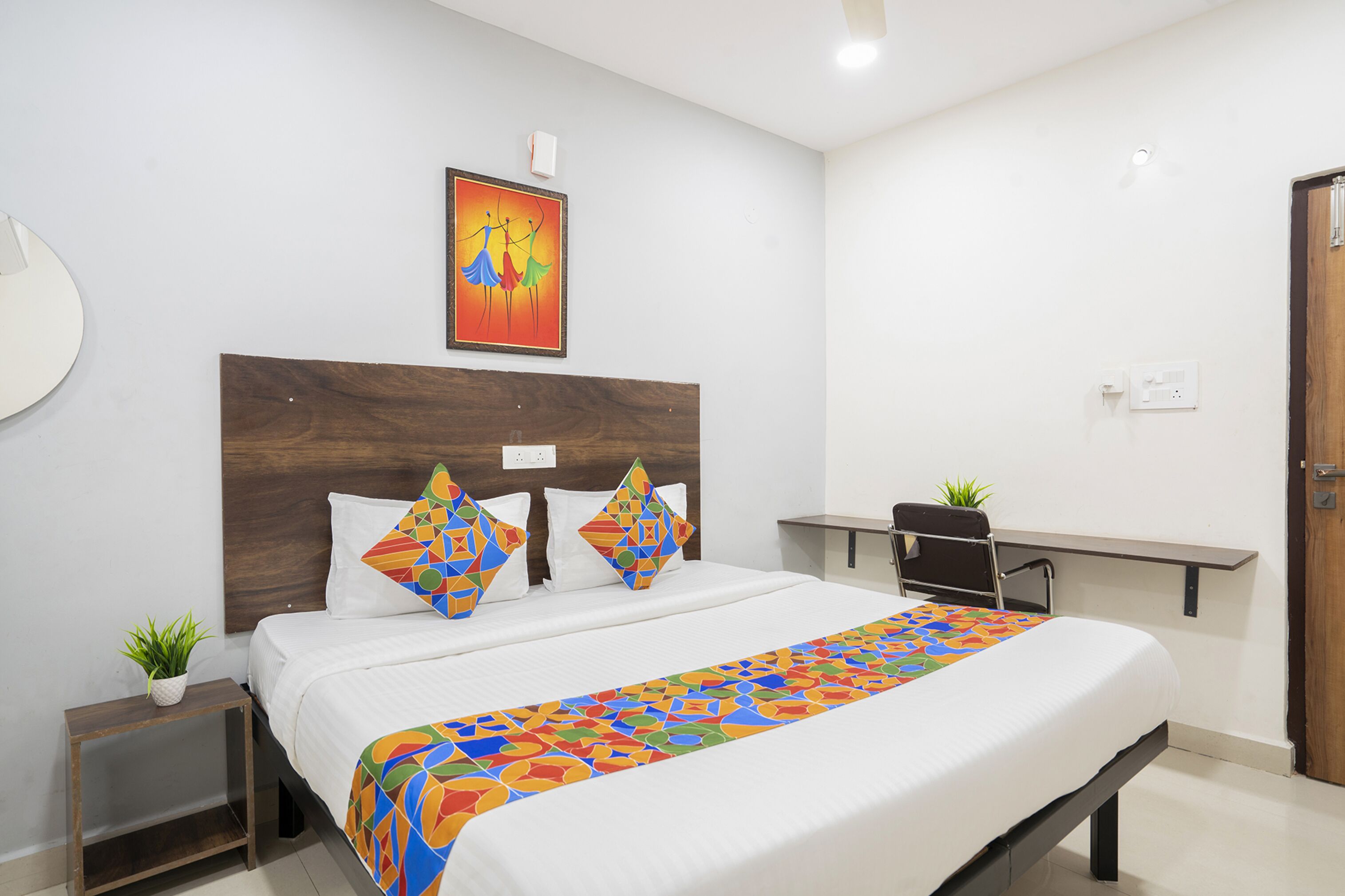 Une chambre d’hôtel bien aménagée, avec un grand lit, une tête de lit en bois, une couvre-lit colorée, une petite table de chevet agrémentée d’une plante et un bureau avec une chaise.