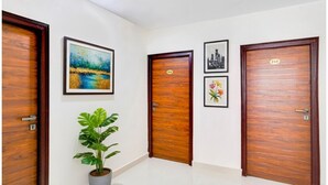 A hallway with three wooden doors, each with a number plate. There are framed artworks on the walls, and a potted plant is visible. - FabHotel Grand Aarvi Prime - Nr. Hafeezpet, Madeenaguda (Hyderabad)