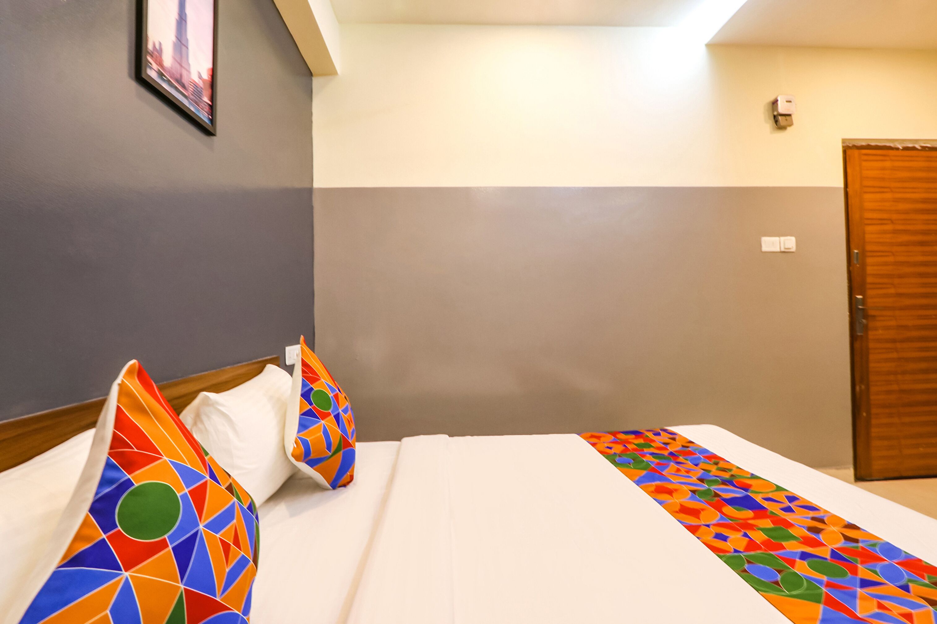 A hotel room with a bed, pillows, a wooden door, and a wall-mounted light switch.