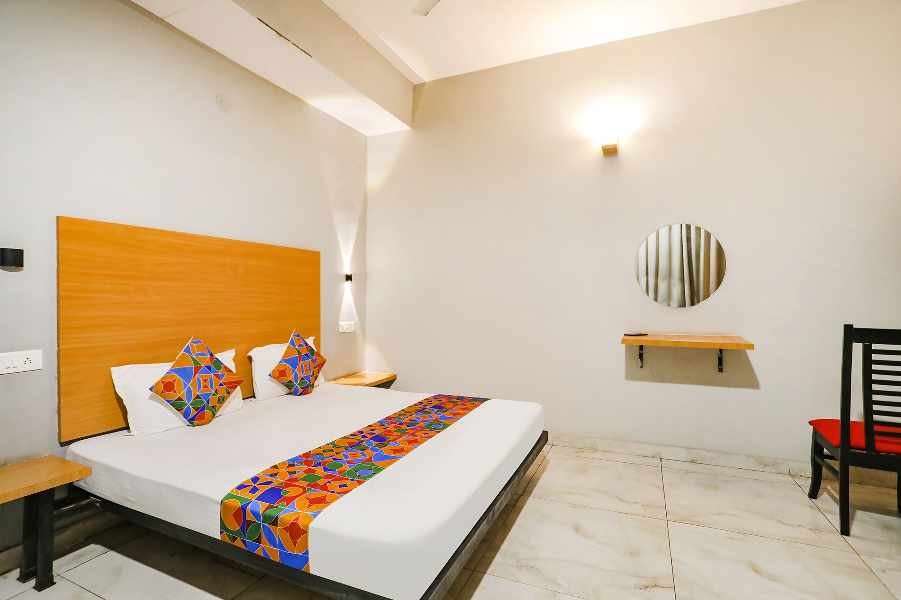 A modern hotel room with a wooden headboard, a colorful bed runner, a round mirror, and a red chair.