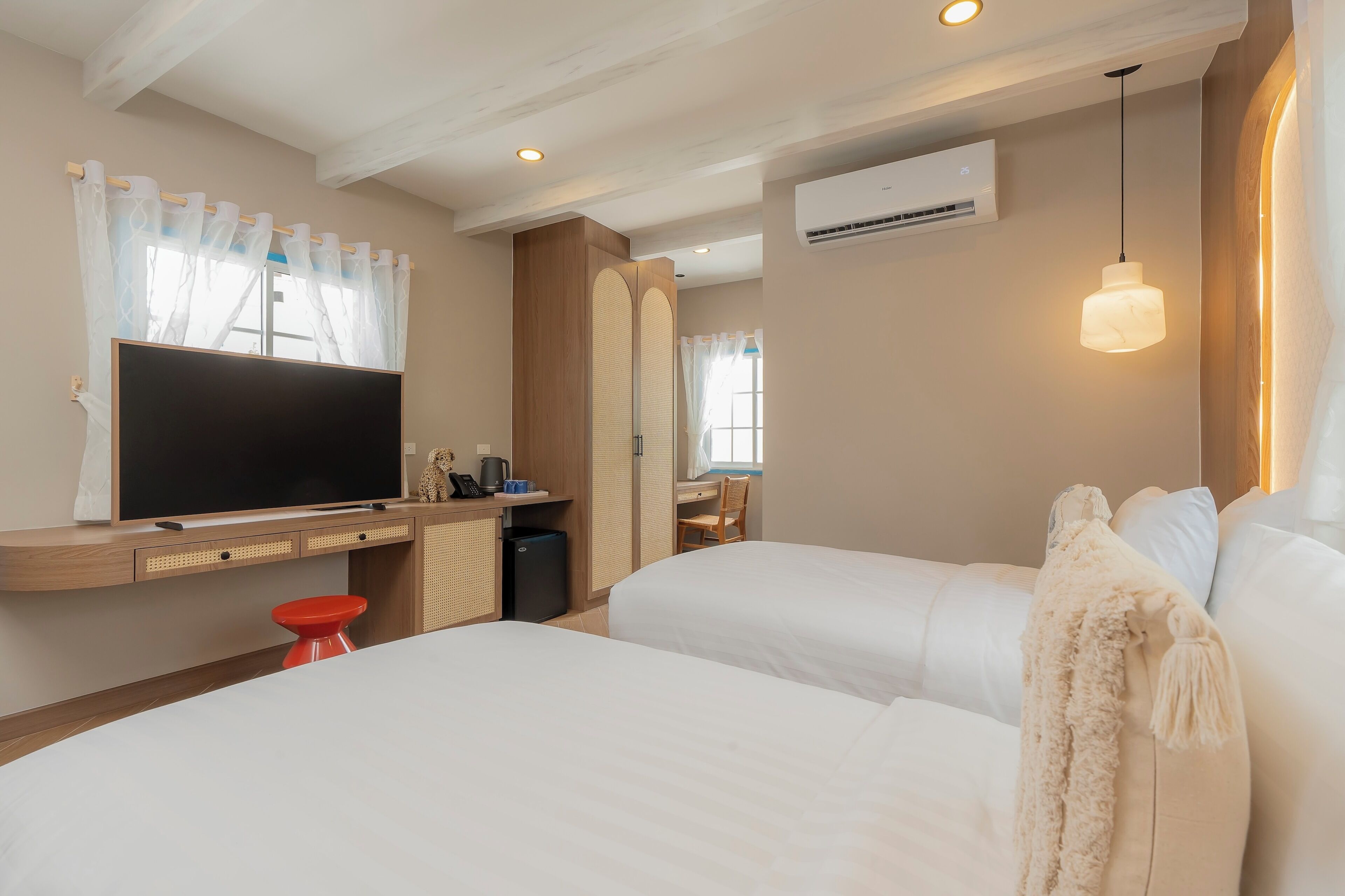 Habitación de hotel con dos camas, un televisor, un escritorio con silla y un taburete rojo.