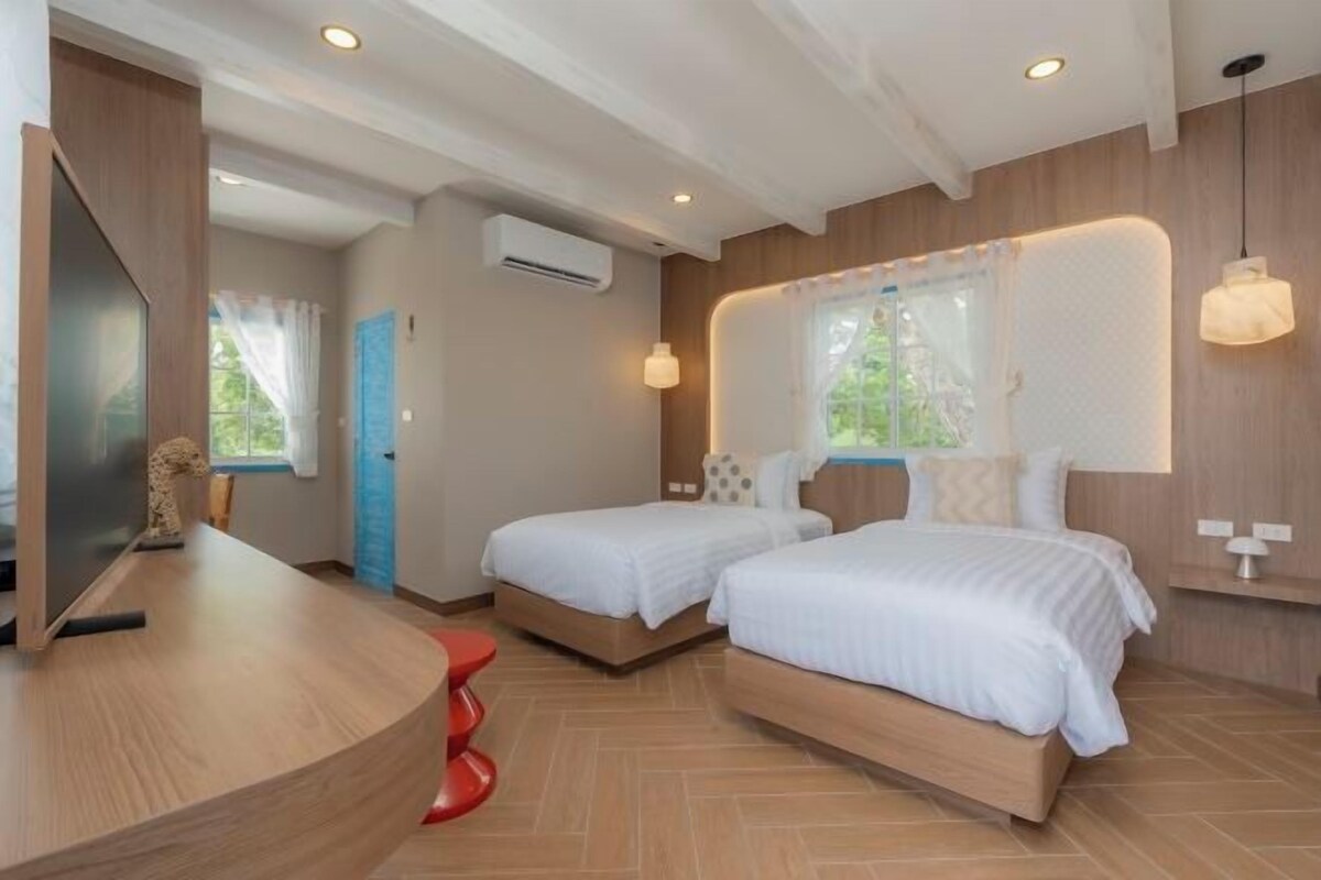 A hotel room with two beds, a curved wooden desk, a red stool, and a large window with curtains.