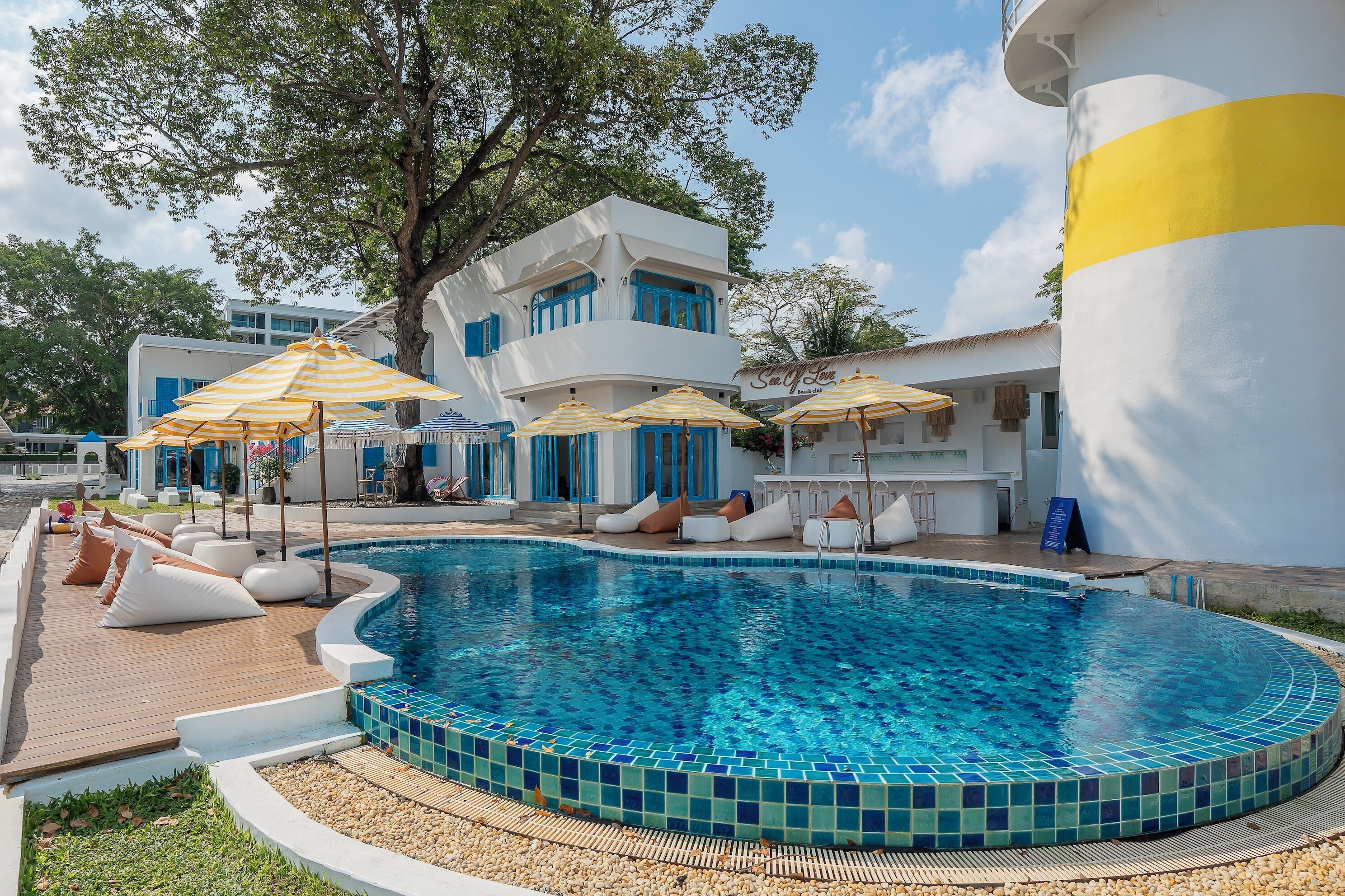 A swimming pool with lounge chairs and umbrellas, a white building with blue accents, and a large tree.