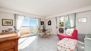 Una habitación con una mesa de comedor, sillas, un sofá rojo, un otoman floral y vistas al mar.