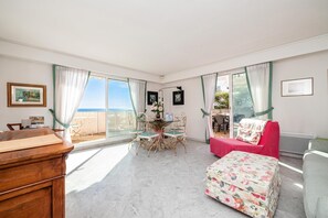 Quarto com mesa de jantar, cadeiras, um sofá vermelho, um banquinho floral e vista para o mar.