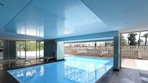 Una piscina interior con paredes de vidrio transparente, una terraza de madera y vistas a un área de estar exterior y palmeras.