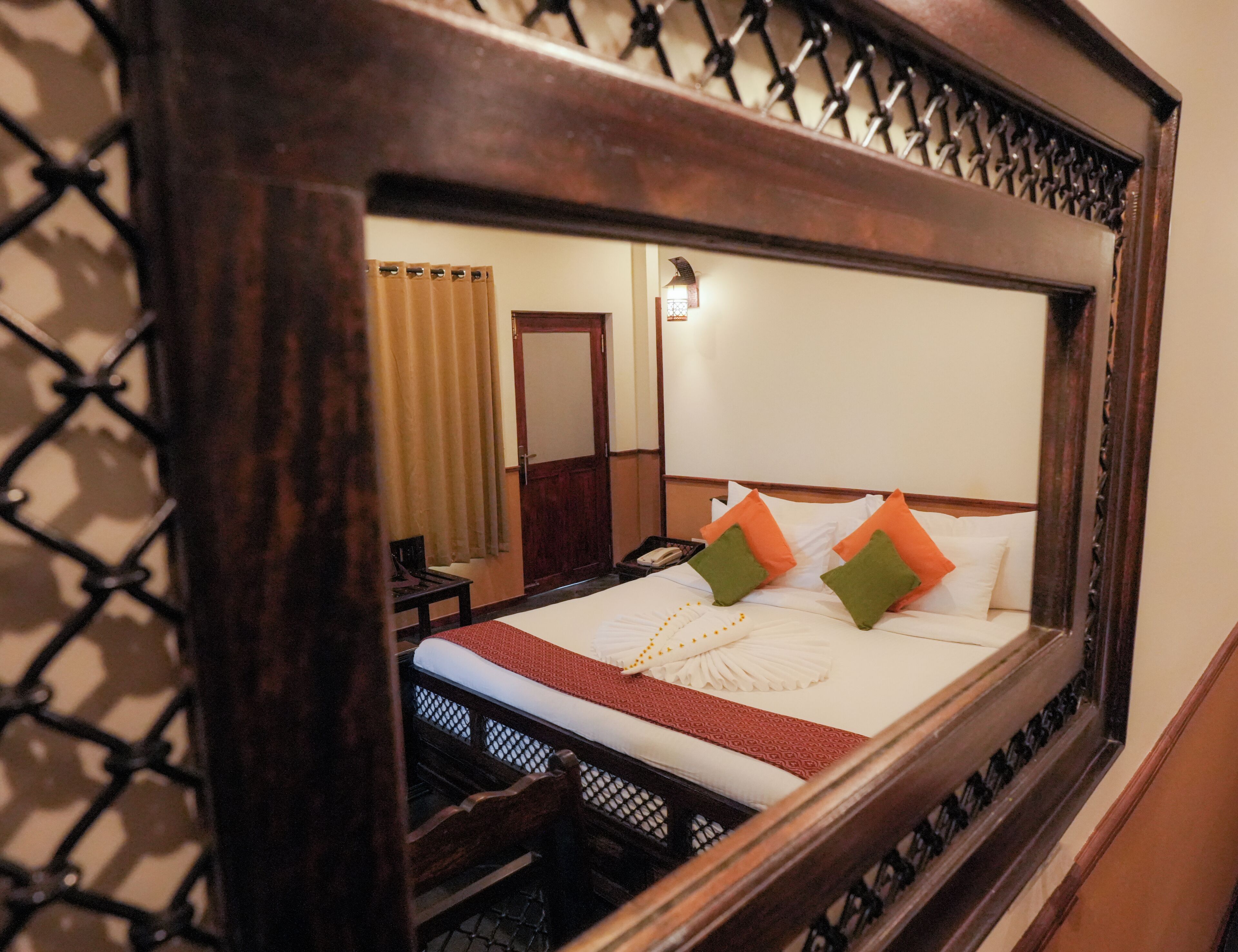A hotel room with a large mirror reflecting a neatly made bed with white linens and orange and green pillows. A wooden headboard and a small table with a chair are visible in the reflection.