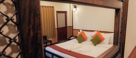 A hotel room with a large mirror reflecting a neatly made bed with white linens and orange and green pillows. A wooden headboard and a small table with a chair are visible in the reflection.
