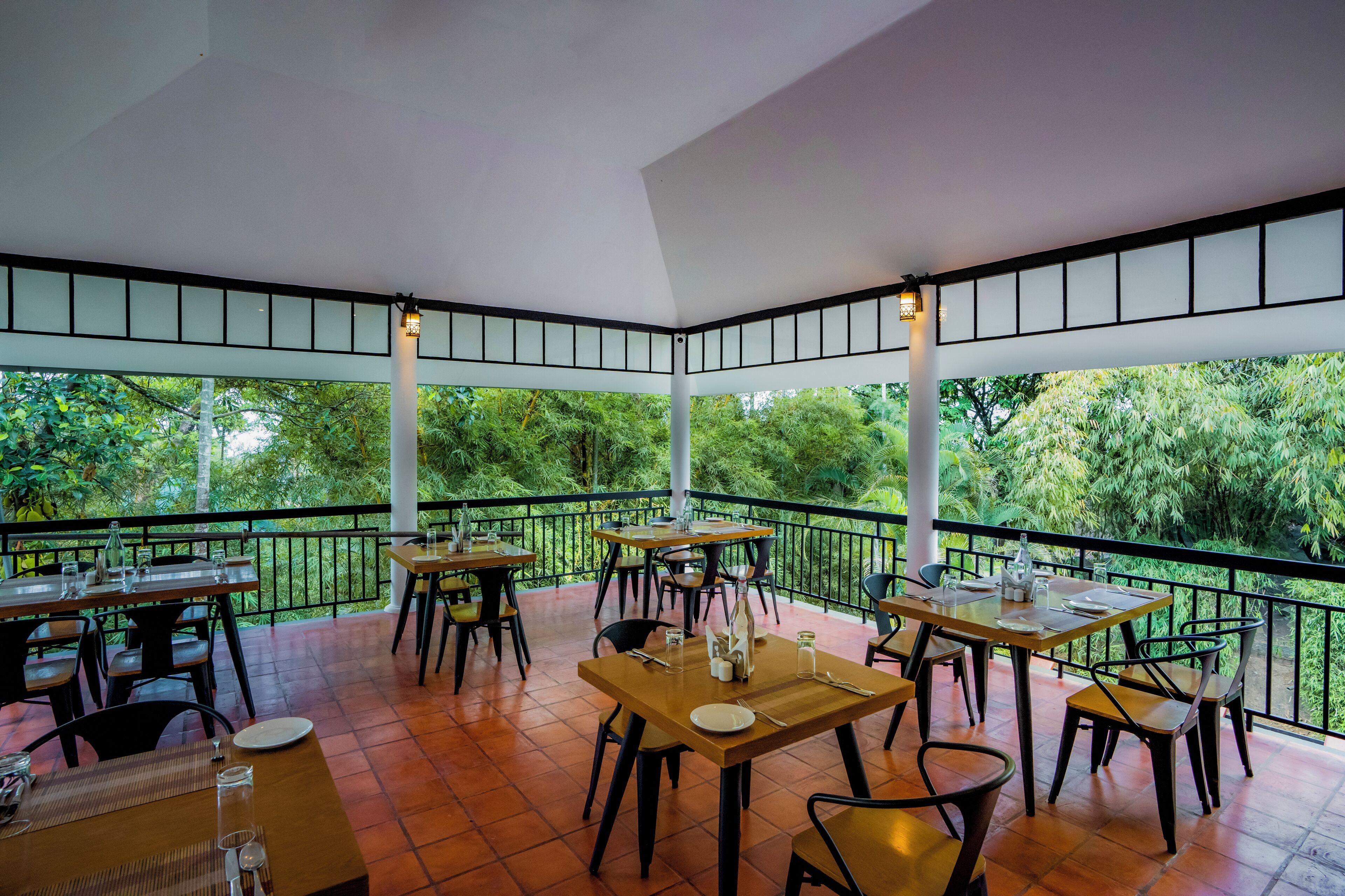 An outdoor dining area with tables and chairs, surrounded by a black railing, and overlooking a lush green landscape.