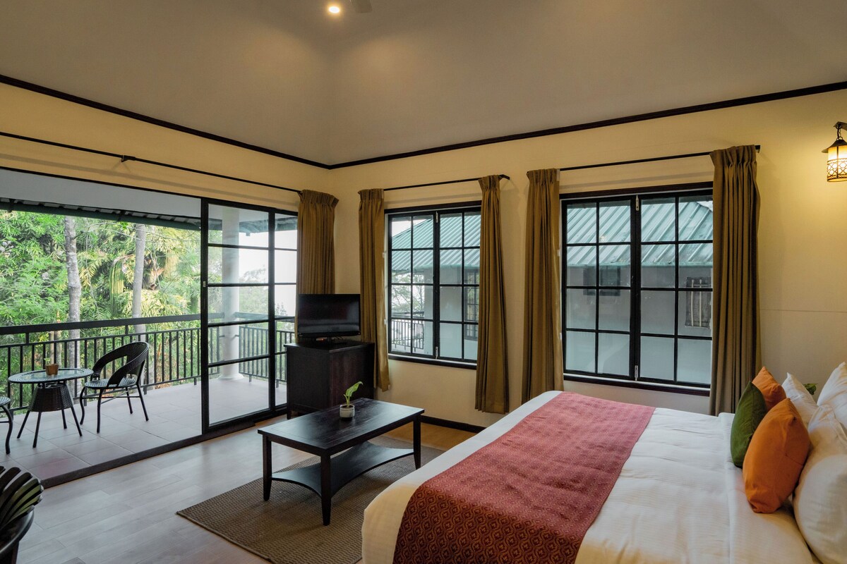 A hotel room with a bed, a TV, a balcony with chairs, and large windows.