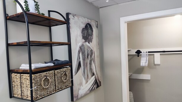 Un baño con estante metálico negro que contiene toallas y una cesta de mimbre, un cuadro de una figura, y un perchero con una toalla colgada.