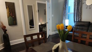 Un comedor con una mesa de madera oscura, sillas y un jarrón con flores amarillas. Hay un cuadro en la pared y una ventana con cortinas.