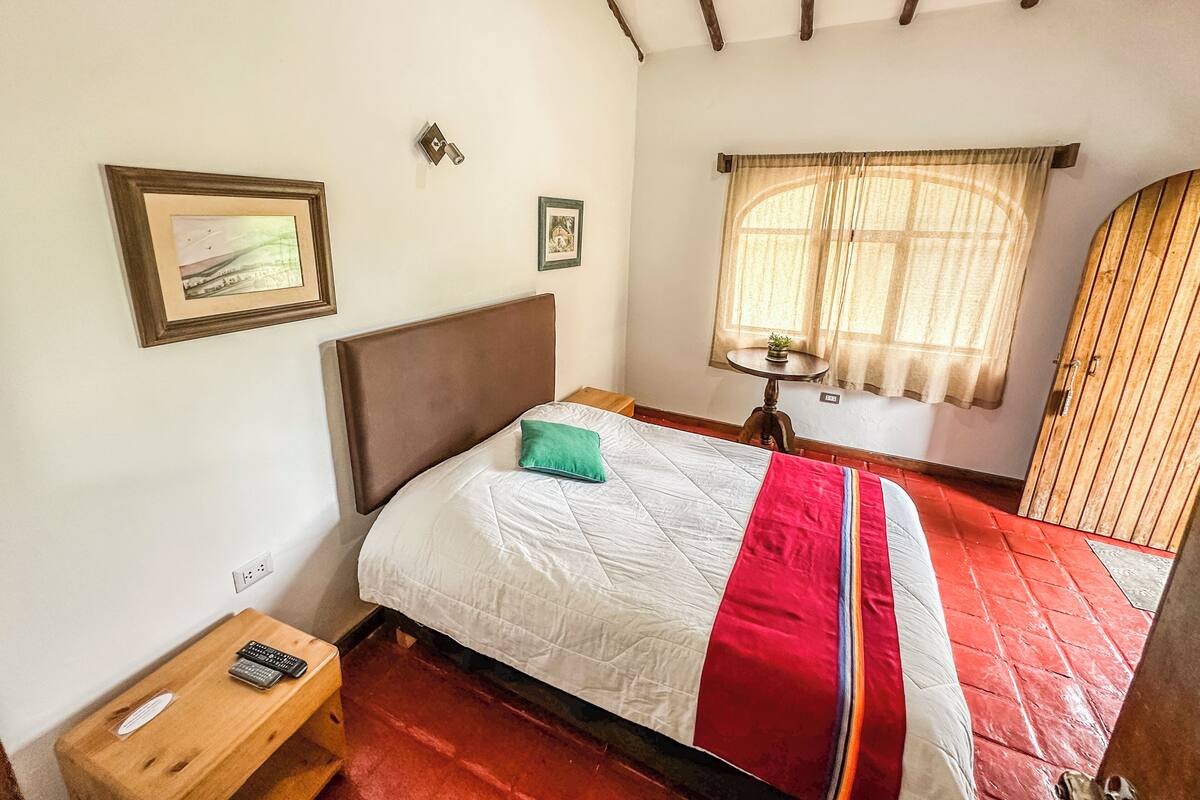 A bedroom with a bed, a wooden nightstand, a small table with a plant, and a window with curtains.