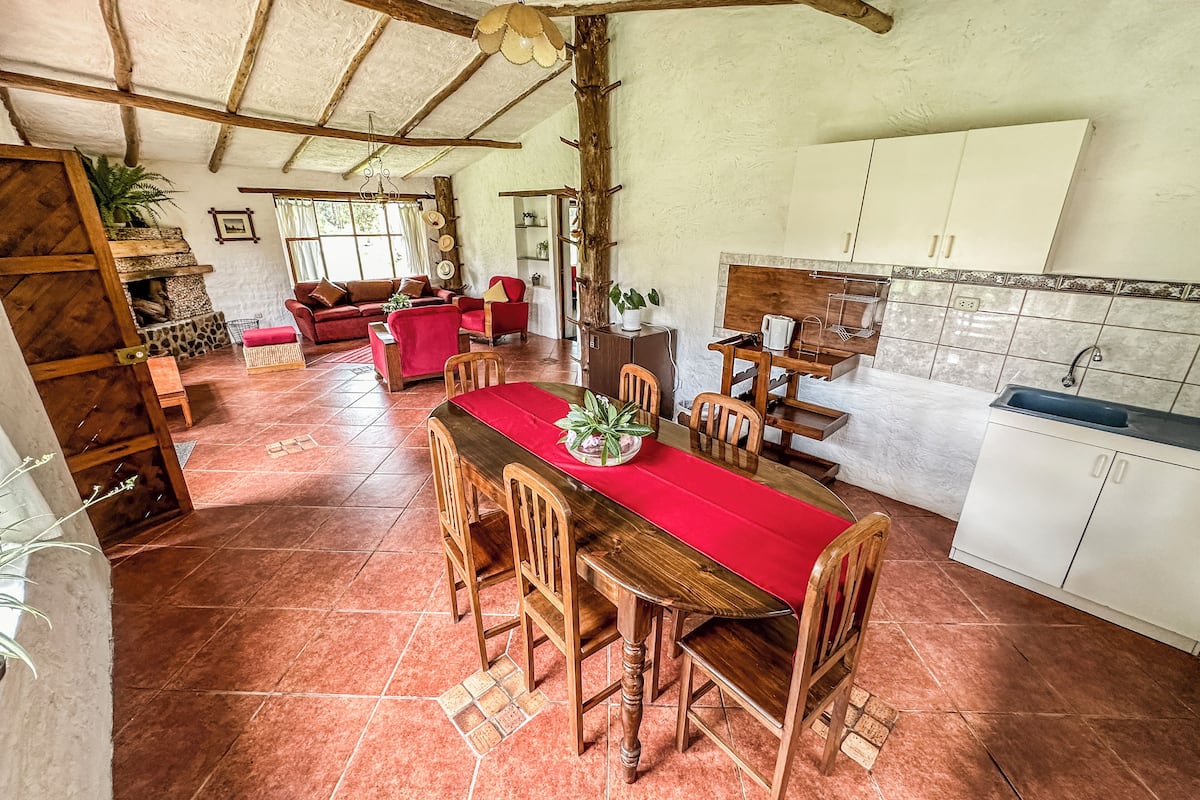 A spacious living area with a red dining table, wooden chairs, and a red sofa. There is a kitchen area with white cabinets and a sink.