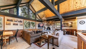 Un salon chaleureux avec une cheminée en pierre, des poutres en bois et une grande fenêtre donnant sur une zone boisée.