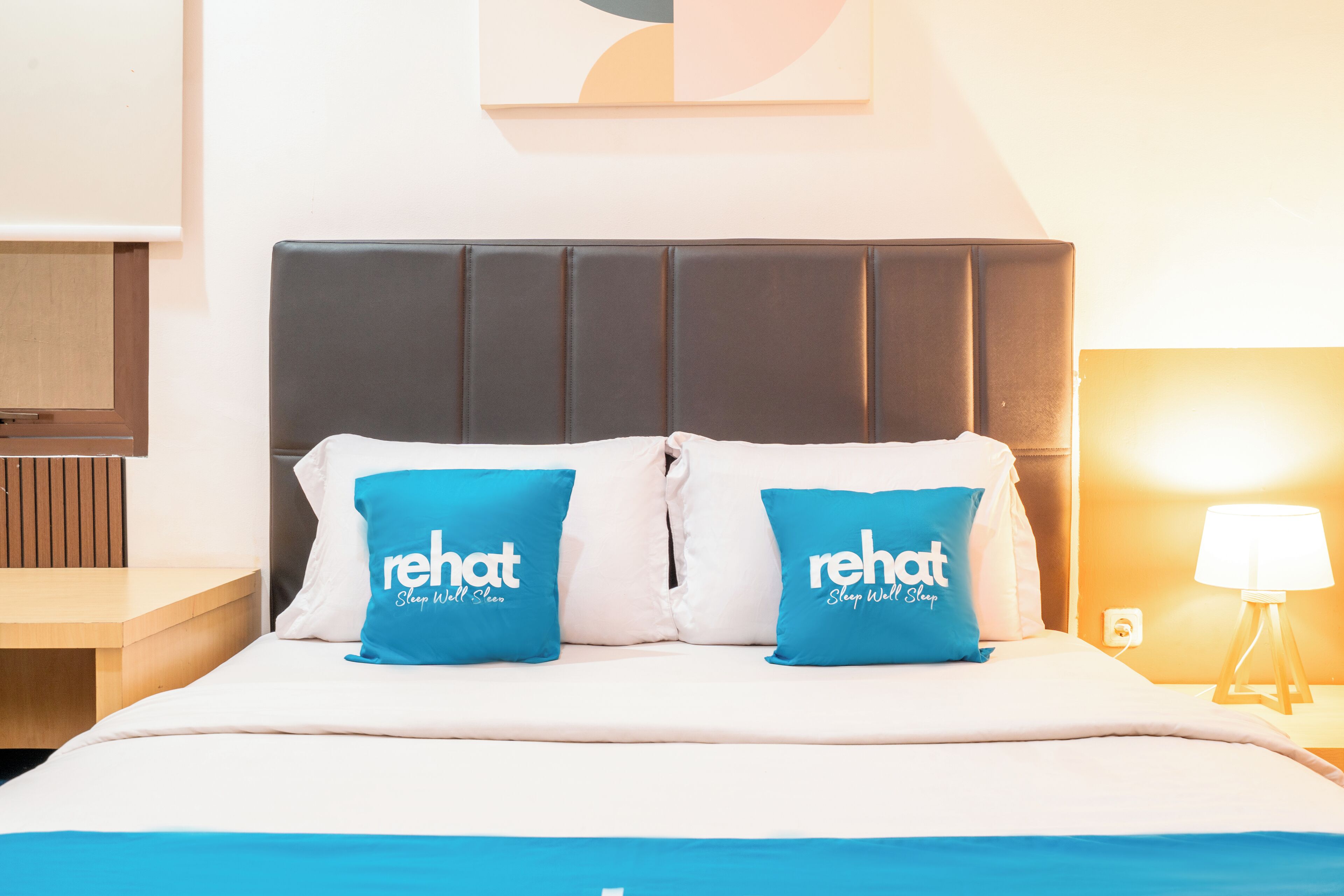 A hotel room with a bed, two blue pillows with 'rehat' branding, a bedside lamp, and a wall-mounted artwork.