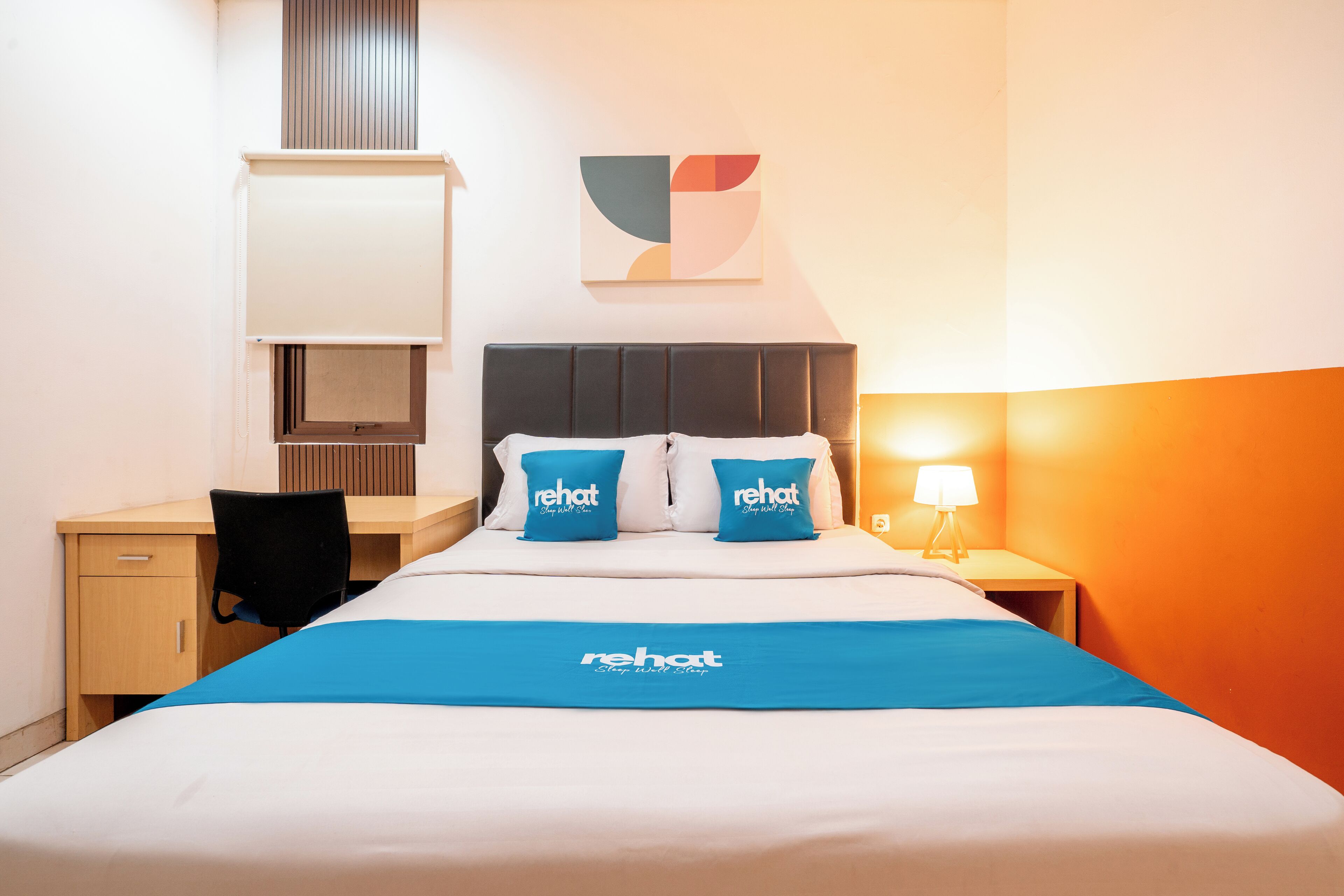 A hotel room with a bed, a desk, a chair, a nightstand, a lamp, and a wall-mounted artwork.