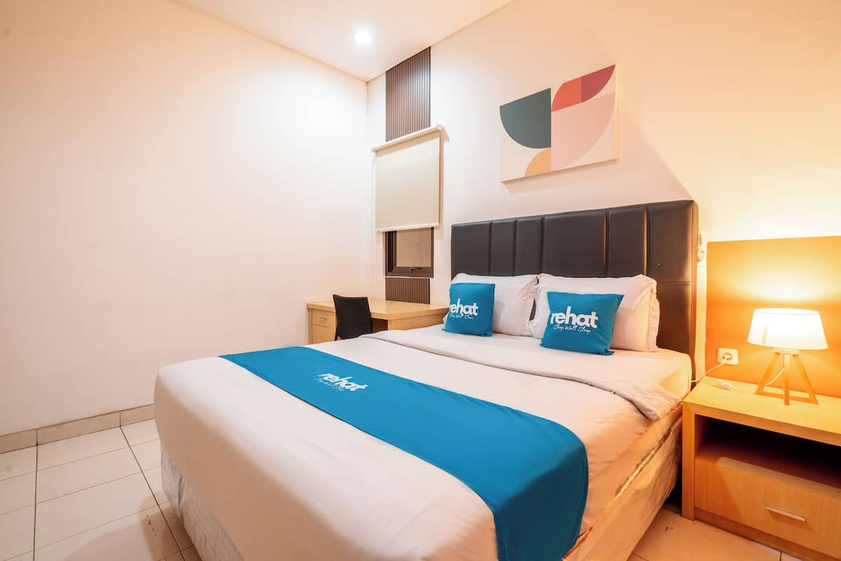 A hotel room with a bed, a desk, a chair, a nightstand, a lamp, and a wall-mounted artwork.