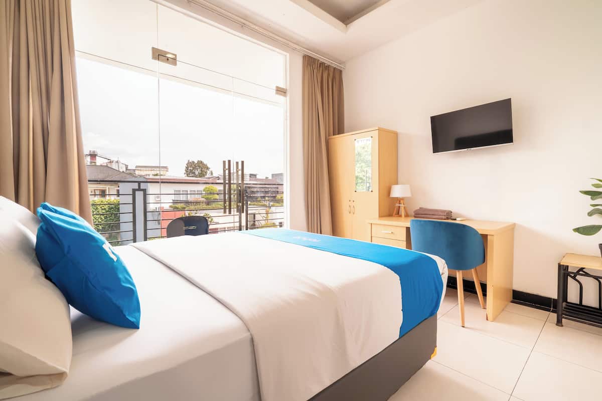 A hotel room with a bed, a desk, a chair, a TV, and a view of the outside.
