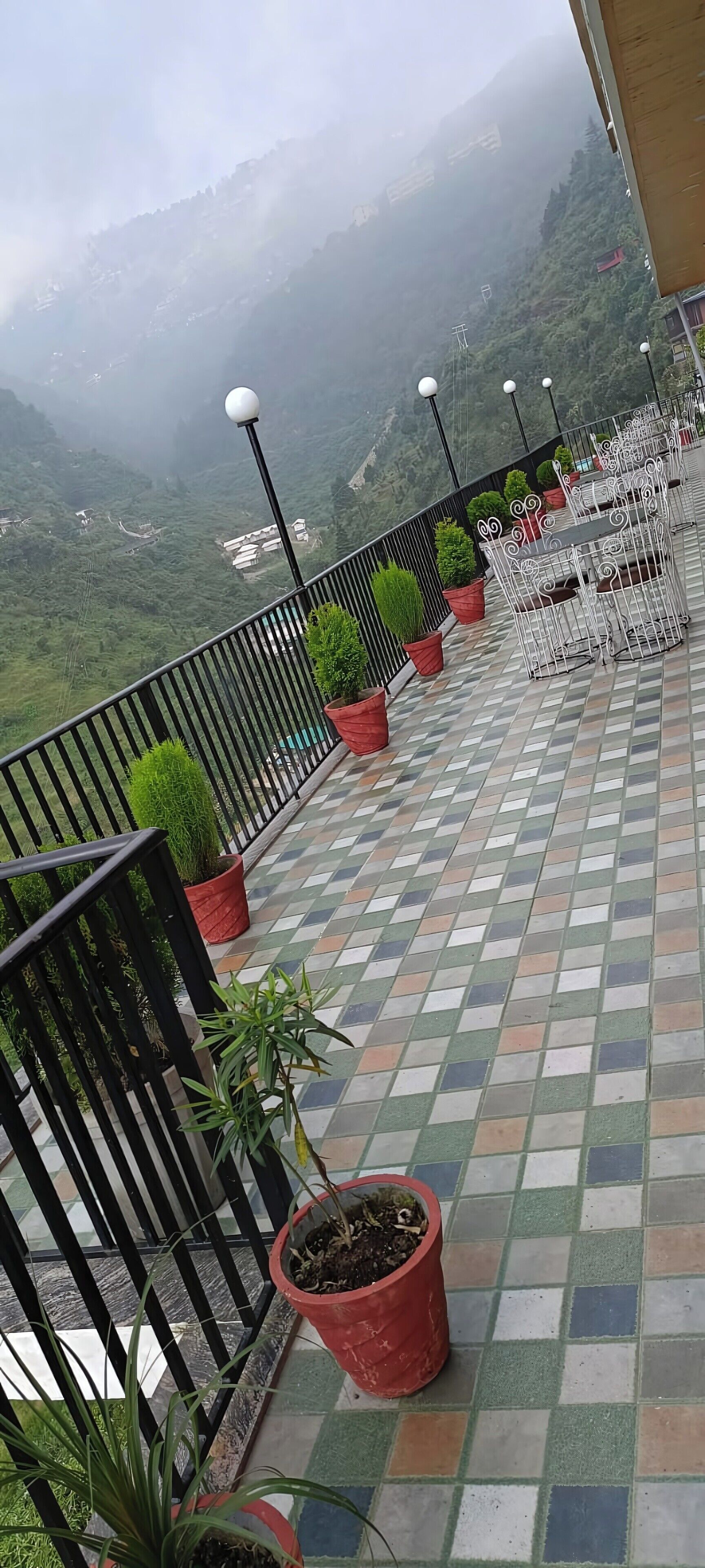 A terrace with a tiled floor, potted plants, and metal railings overlooking a mountainous landscape.