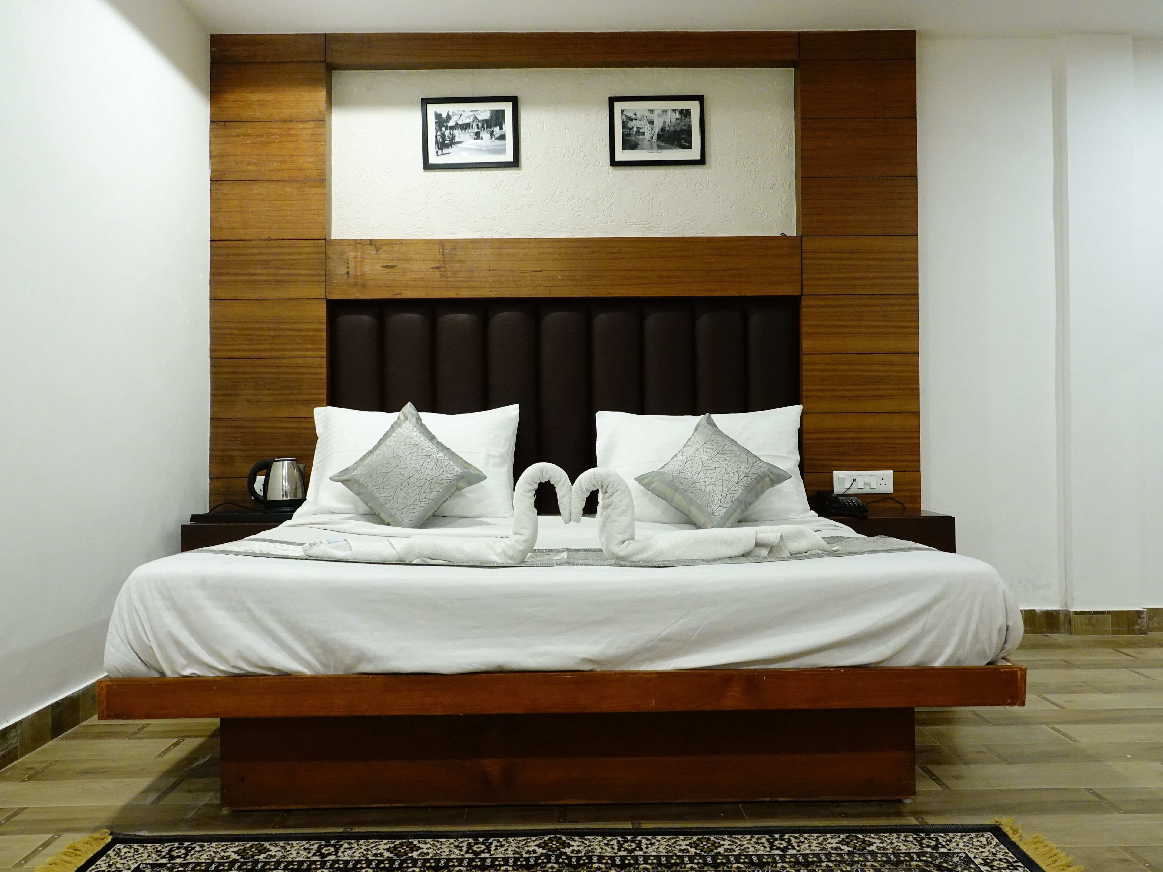 A large bed with a wooden headboard, two pillows, a bedside table with a phone, and a wall-mounted picture frame.