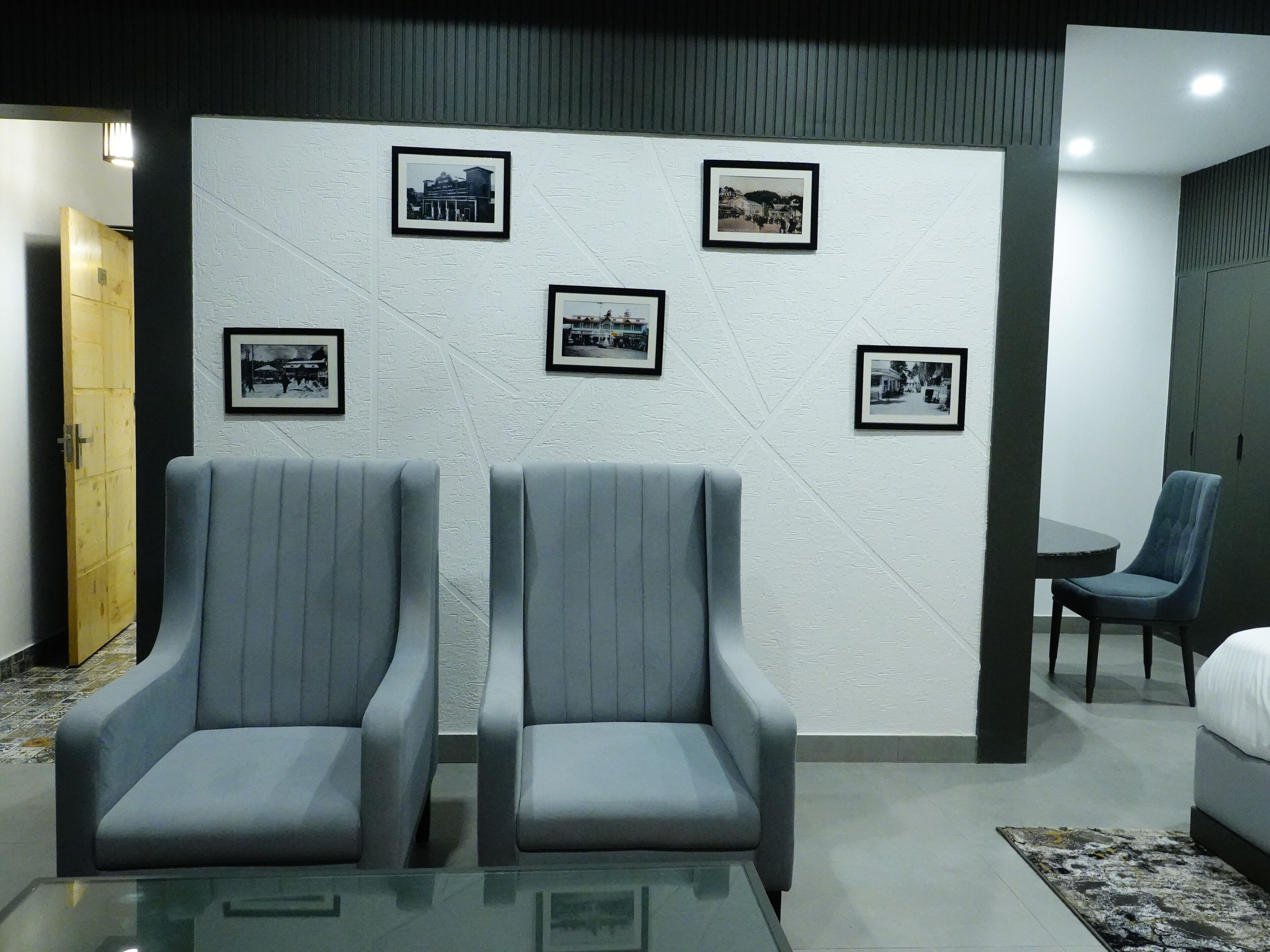 A hotel room with two grey armchairs, a small table, and a bed. The wall features four framed black and white photographs.