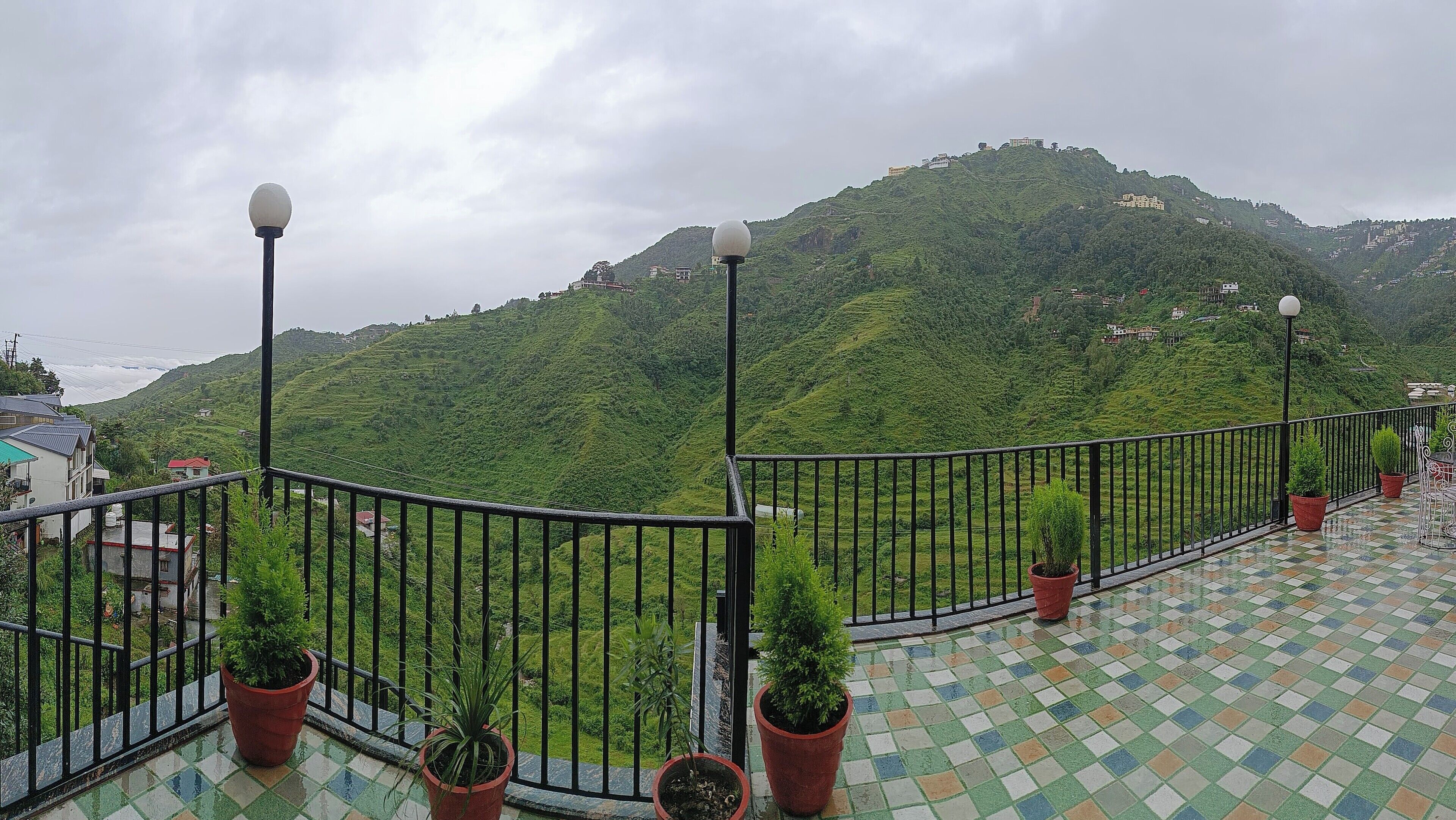 A balcony with a tiled floor, potted plants, and a view of a green hillside with buildings.