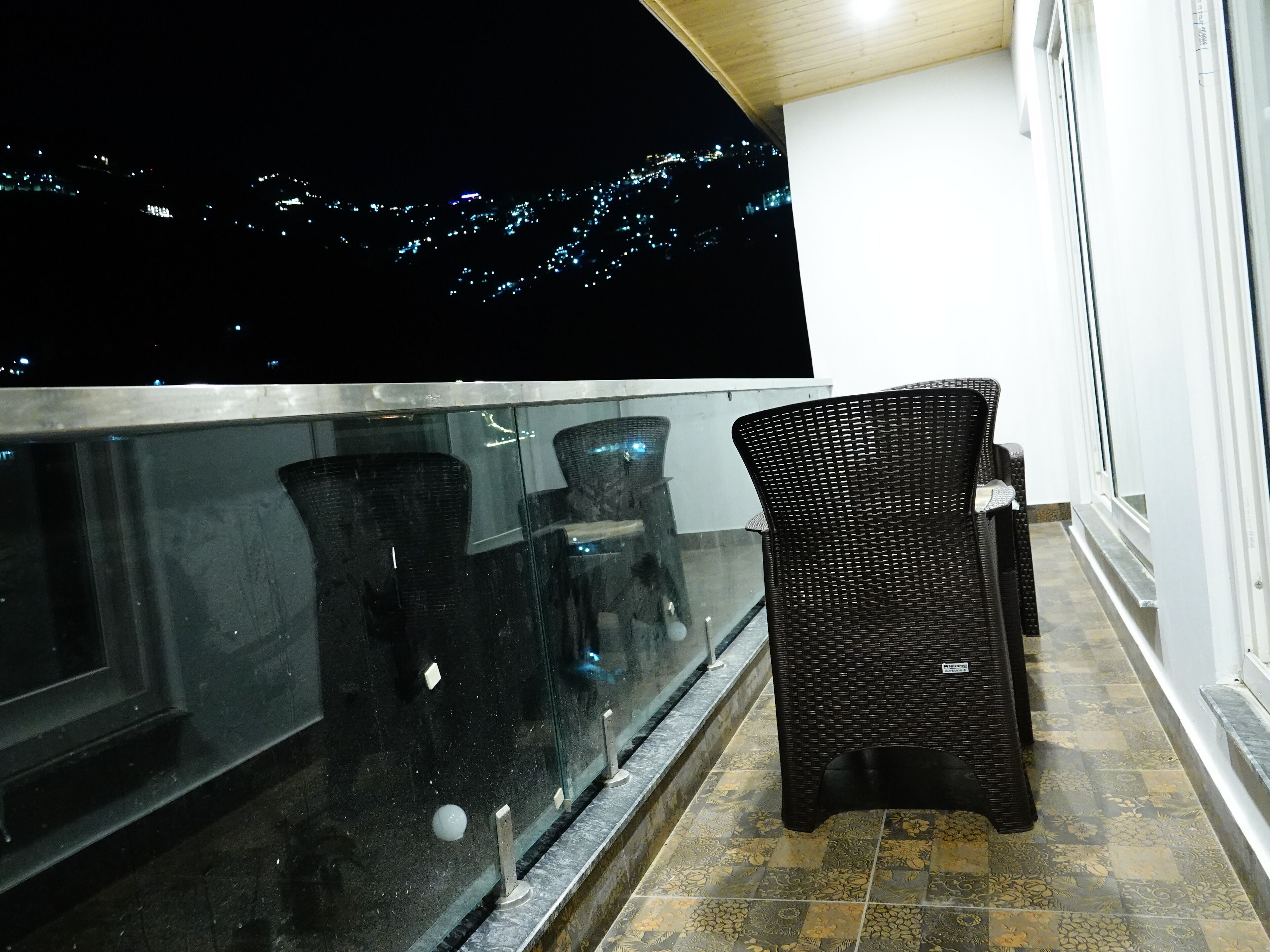 Ein Balkon mit Glasgeländer, einem Korbstuhl und Blick auf die nächtlichen Stadtlichter.