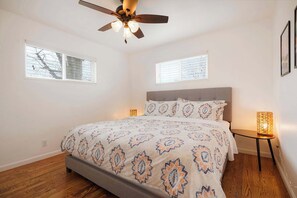 Quarto com cama, ventoinha no teto, duas janelas e mesa de cabeceira com abajur.