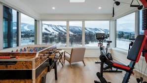 A spacious room with a foosball table, exercise equipment, and large windows offering a view of snow-covered mountains. - ULTRA LUXURY BLUE ROSE CHALET BUILT IN 2023 HOSTS 12 GUESTS GYM/HOTTUB/GAMES  (blue mountain)