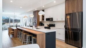 A modern kitchen with a central island, bar stools, a stainless steel refrigerator, and pendant lighting. - ULTRA LUXURY BLUE ROSE CHALET BUILT IN 2023 HOSTS 12 GUESTS GYM/HOTTUB/GAMES  (blue mountain)