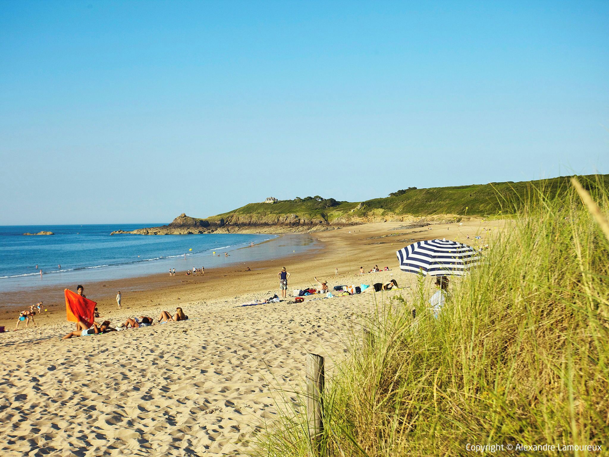 A sandy beach with people sunbathing and walking, a grassy area with a striped umbrella, and a house on a hill in the background.