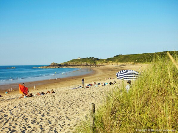 Une plage de sable où des gens se prélassent au soleil et se promènent, une zone herbeuse avec un parasol rayé, et une maison située sur une colline en arrière-plan.