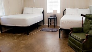 A room with two beds, a green armchair, a small table with a lamp, and a window with blinds.