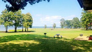 A grassy park area with picnic tables and a mailbox, overlooking a body of water and trees.