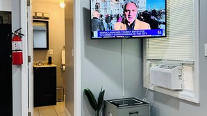 A living room with a television displaying a news program, a potted plant, a small cabinet, and a window with blinds.