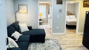 A modern living room with a sectional sofa, a patterned rug, and a hallway leading to a bedroom with a bed and a nightstand.