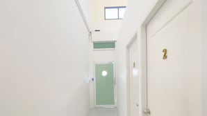 Un pasillo estrecho, con paredes blancas, una puerta verde al final, una pequeña ventana en lo alto y una puerta con el número 2.