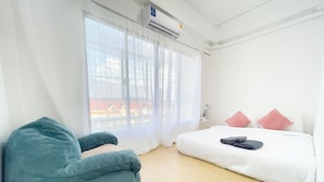 Una habitación de hotel con una cama, una silla azul, una ventana con cortinas translúcidas y un aire acondicionado de pared.