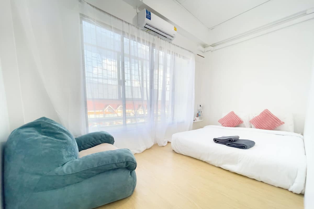Een hotelkamer met een bed, een blauwe stoel, een raam met dunne gordijnen en een wandairconditioning.