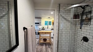 Un pasillo estrecho con una puerta de ducha de vidrio, una mesa de madera con un cuenco, y una cocina al fondo.
