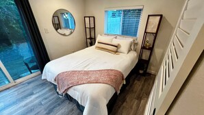 Un dormitorio moderno con una cama grande, un espejo redondo, una ventana con persianas y vistas al exterior con vegetación.