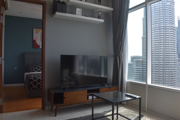 A modern living room with a large flat-screen TV, a glass coffee table, and a city view through a large window.
