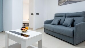 Un salon moderne avec un canapé gris, une table basse blanche ornée de fruits et un tableau représentant une route accroché au mur.